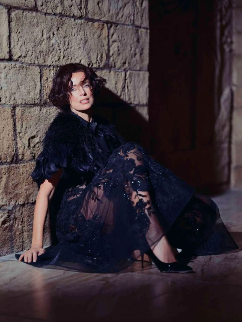A woman poses in an elegant black dress, seated against a textured stone wall, with soft lighting and a glamorous fashion style.
