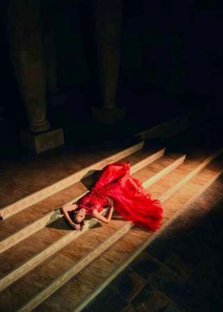 Woman in red dress lies on marble steps with triangle pattern, bathed in a spotlight, with dark stone columns in background.