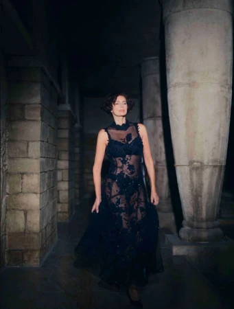 Heather Stewart-Whyte in a dark sheer embroidered dress walking through a stone hall with columns for Glamour Bulgaria.