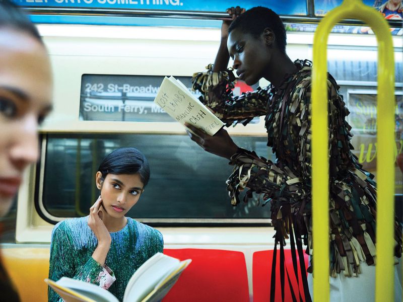 Three women on subway train reading books. Two are wearing patterned tops. Subway car interior with seats and window visible.