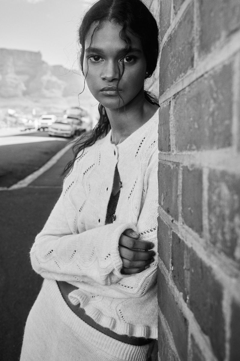 Portrait of a young woman with arms crossed, leaning against a brick wall in a black and white photo with mountains in the background.