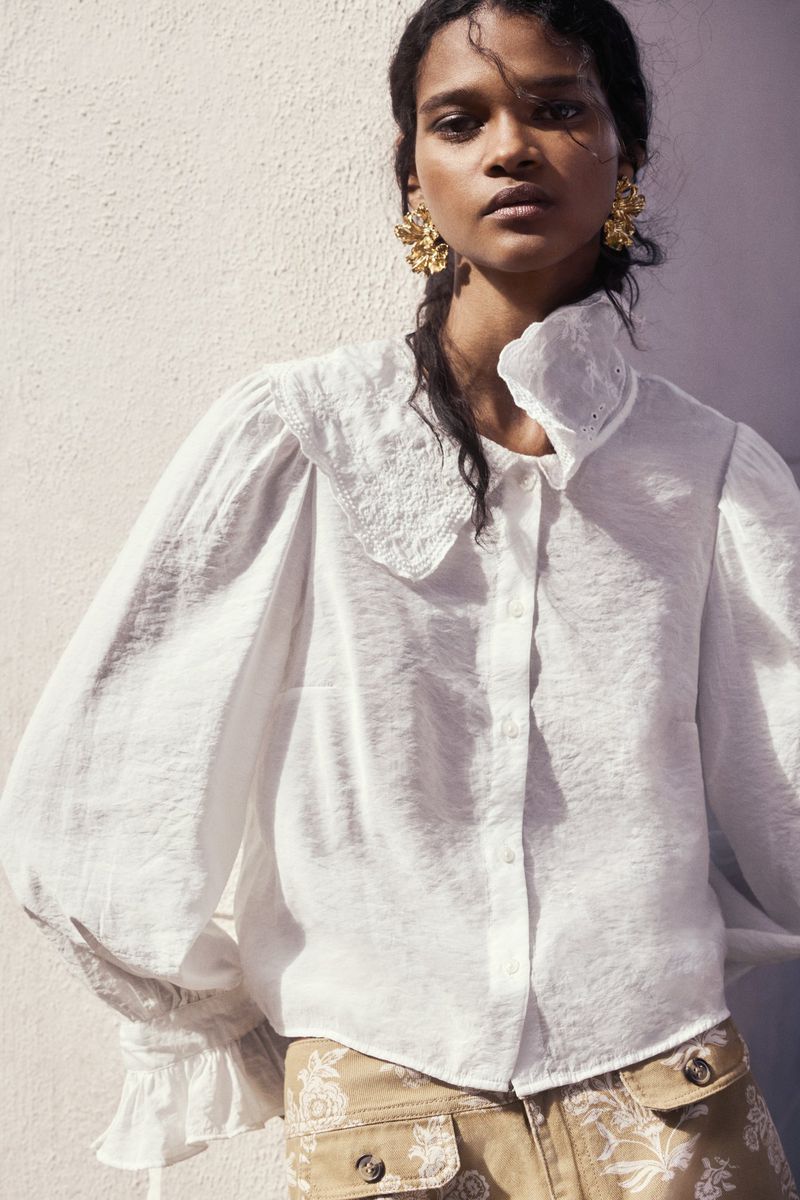 A woman in a white blouse with large collar and tan pants with floral design poses in front of a wall with soft, natural lighting.