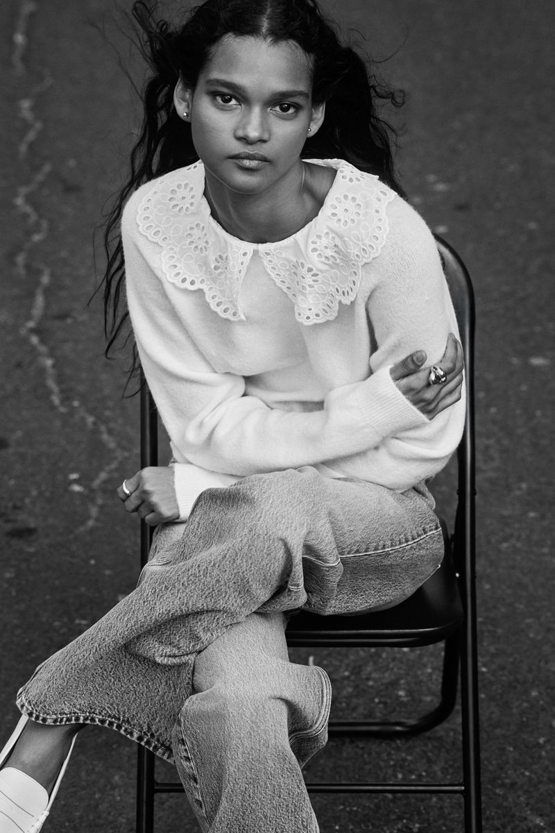 Black and white portrait of a young adult female sitting on a chair wearing a sweater with a decorative collar and wide-leg jeans.