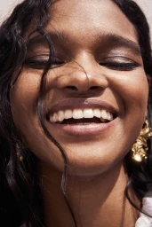 Close-up portrait of smiling woman wearing gold earrings with dark hair blowing across face for H&M Cape Town campaign