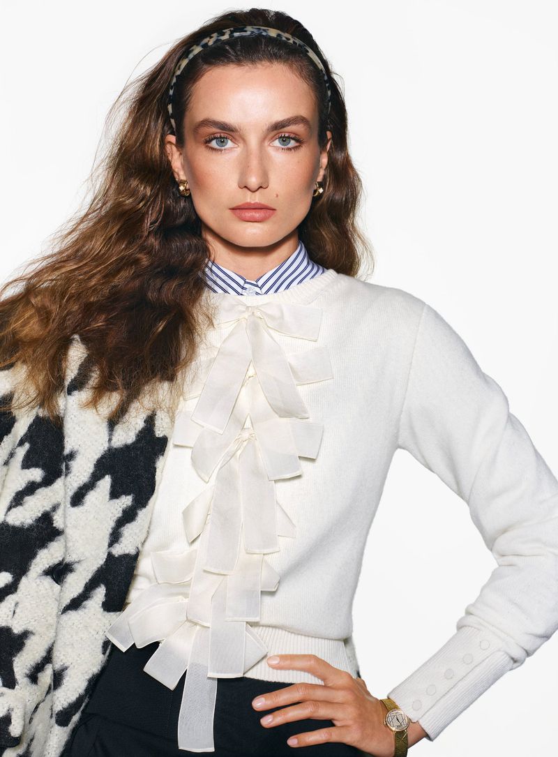 Portrait of a stylish adult woman with long brown hair wearing a cream-colored sweater, black pants, and a houndstooth jacket.