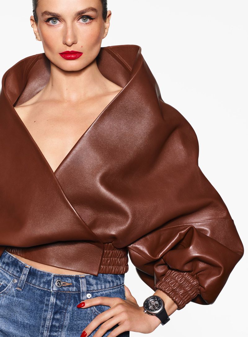 Close-up of a woman with red lipstick wearing a brown leather jacket and blue jeans, posing in a studio with a white background.