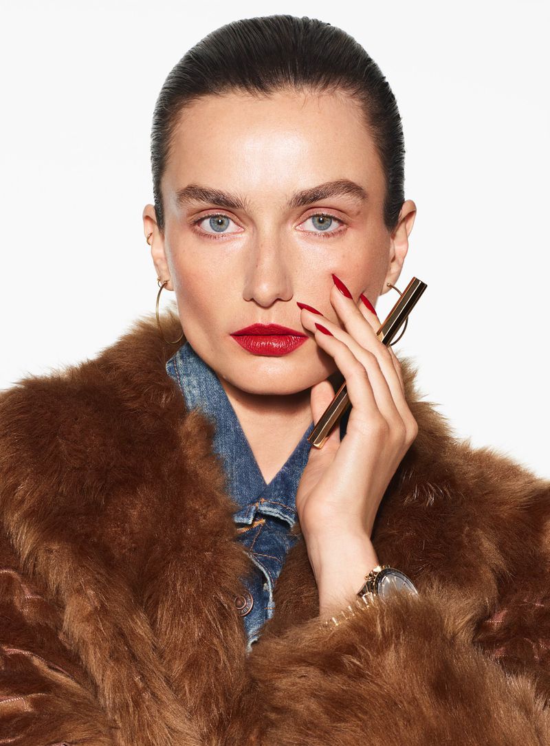 Close-up portrait of a woman with red lipstick and nails wearing a fur coat and holding a gold object, against a white background.