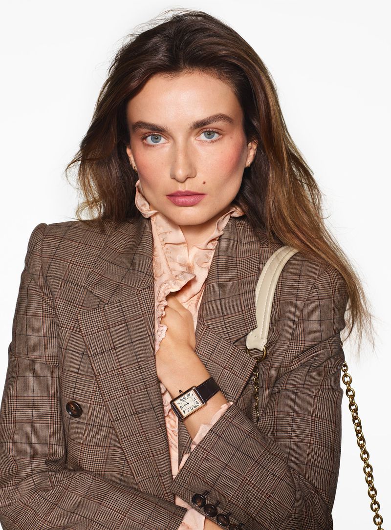 Elegant woman in a plaid blazer and ruffled blouse, showcasing a wristwatch. The model is posed against a bright white backdrop.