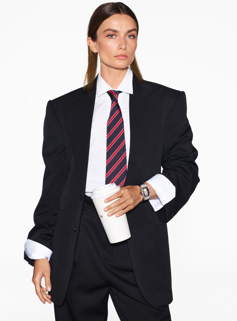 Brunette woman in an oversized black blazer, white shirt, and red-striped tie, holding a coffee cup against a clean white background.