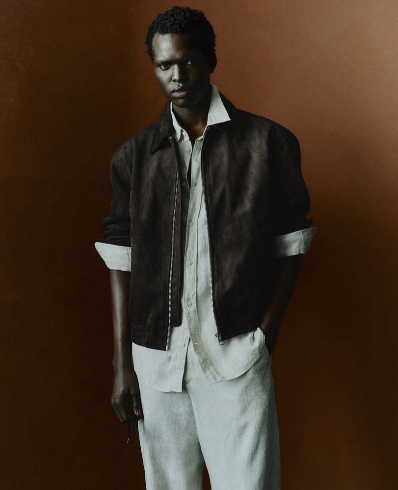 Portrait of a Black male model wearing a dark brown collared jacket and light grey shirt and pants against a brown background.
