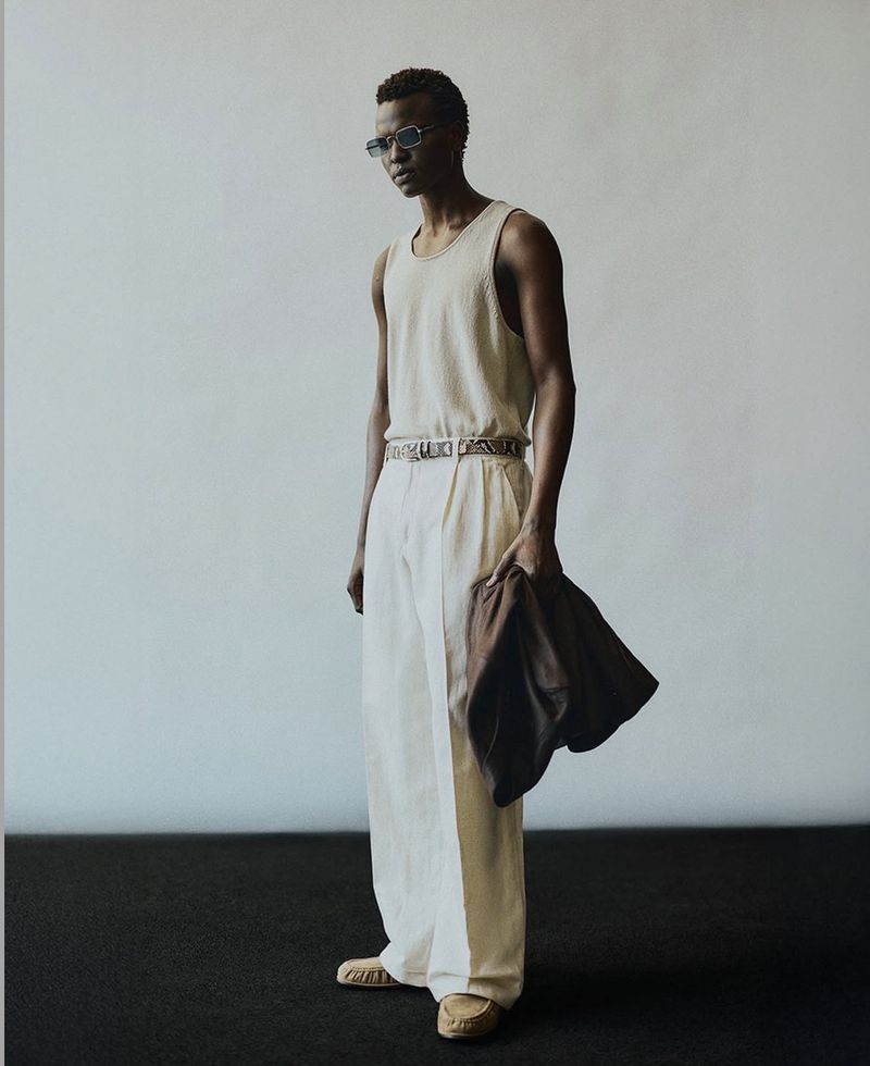 Full-length shot of a Black male model in a tank top and white linen pants, holding a brown jacket in a studio with a black floor.