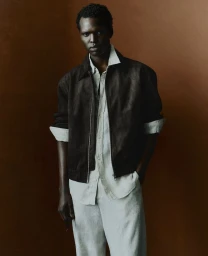 Model wearing a dark brown jacket, light shirt, and trousers against a warm textured brown backdrop.