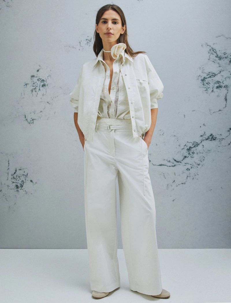 A young adult woman poses in wide-leg ivory pants, ivory blouse and jacket in a full-length studio fashion shot against a mottled background.