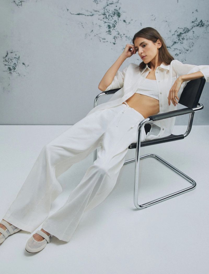 A young woman in a white linen outfit and ballet flats poses on a modern chair in a photo studio, with a marbled wall backdrop.