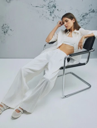 A young woman in a white linen outfit and ballet flats poses on a modern chair in a photo studio, with a marbled wall backdrop.