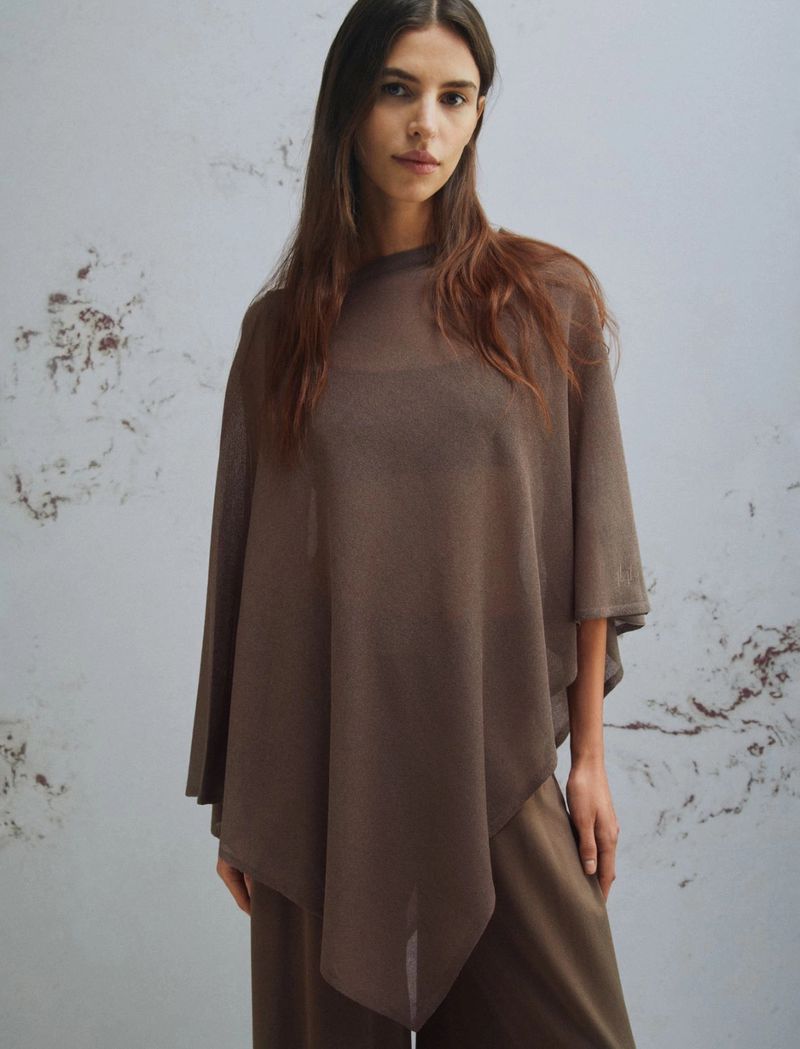 Woman in a brown poncho and pants against a textured background. Fashion shot featuring neutral tones and soft lighting. Casual wear.