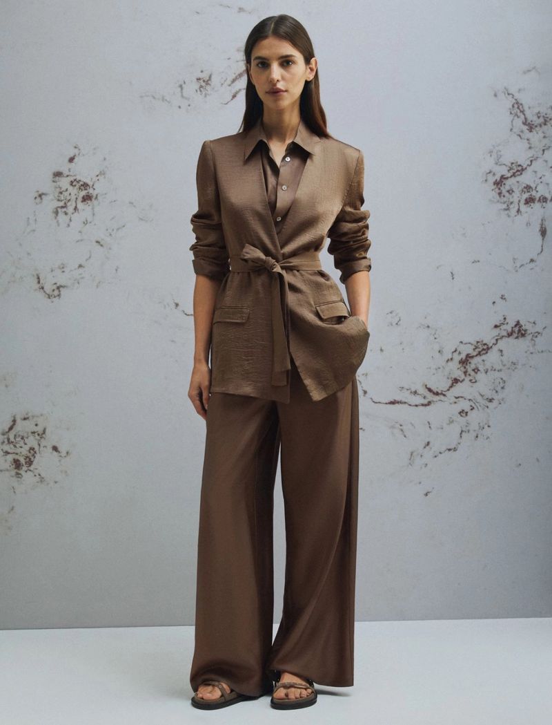A woman models a brown tailored jacket and wide-leg pants, accessorized with strappy sandals, in a full shot against a mottled background.