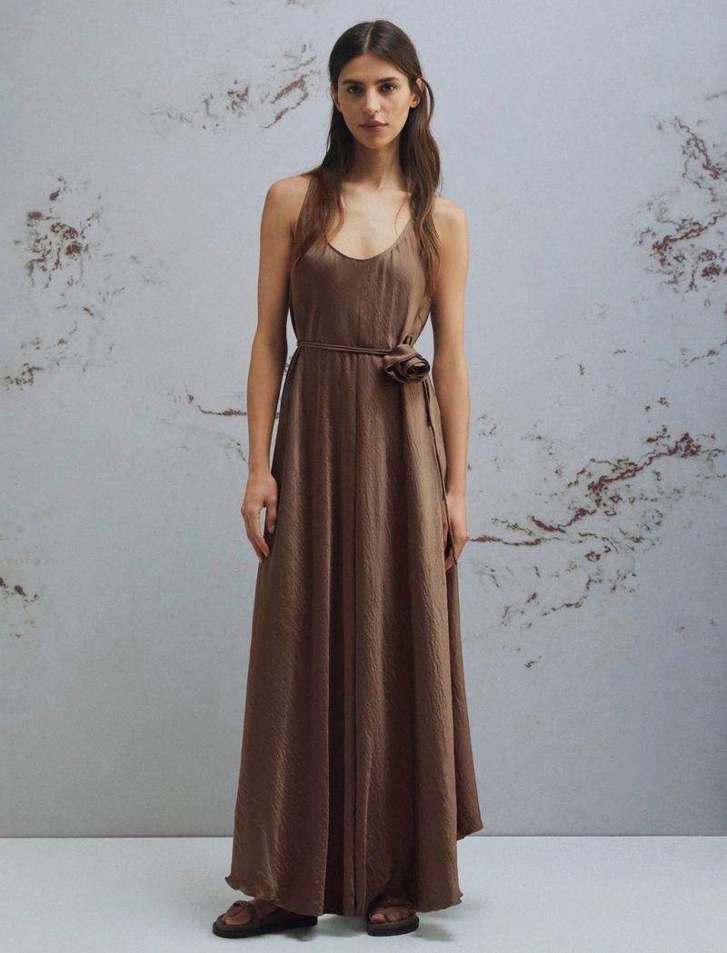 A full shot of a slender woman with long brown hair modeling a long, earth-toned dress with a flower detail against a mottled backdrop.