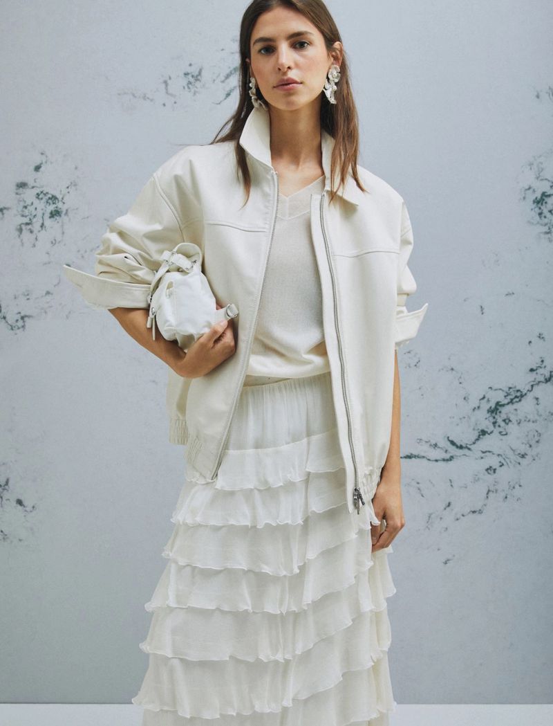A woman in an off-white bomber jacket, matching sweater, and layered frilly skirt poses indoors against a mottled grey background.
