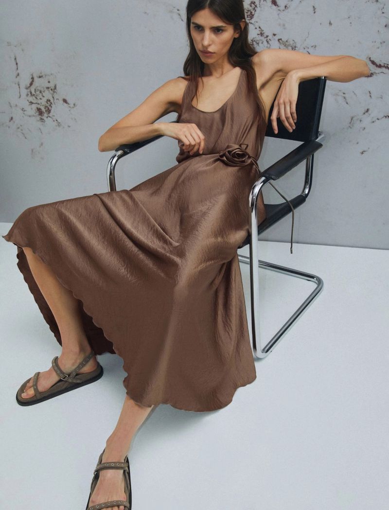A woman sits in a modern chair, wearing a brown slip dress and strappy sandals in a minimalistic studio setting with grey backdrop.