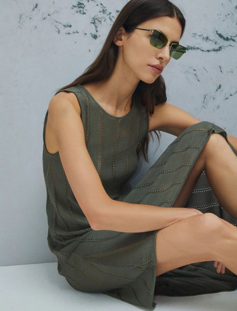 Brunette woman with green sunglasses wearing an olive green dress, sitting indoors against a neutral wall with mottled texture.