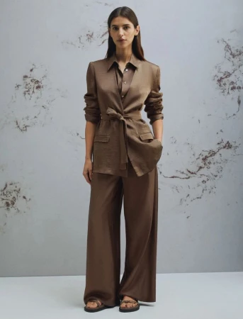 Model standing against a patterned wall wearing a brown belted jacket and matching wide-leg trousers