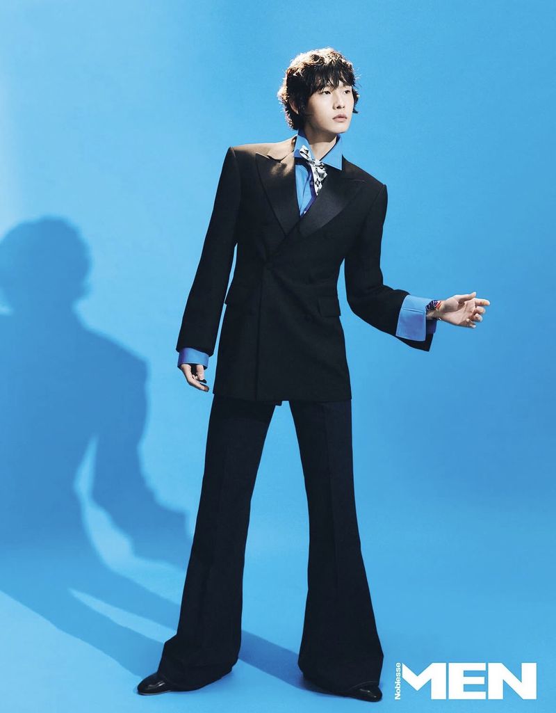 Full-length portrait of a slender adult male with dark hair posing in a black suit and flared pants against a blue backdrop.