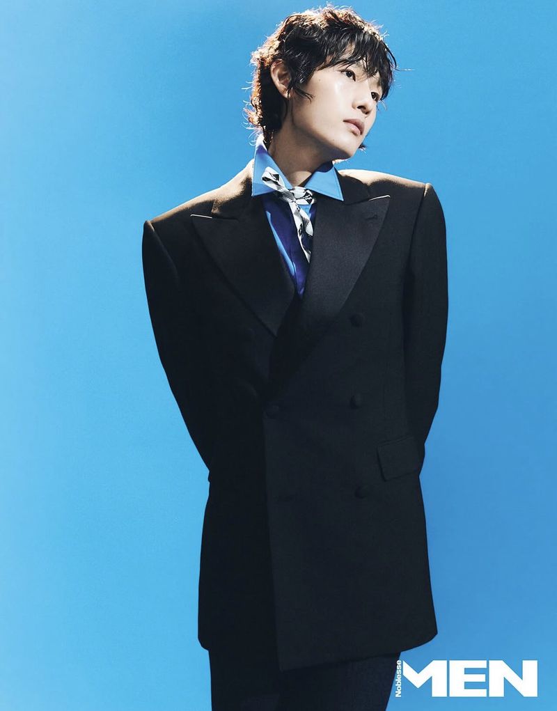 Portrait of a young adult in a black suit jacket, blue shirt, and patterned necktie, against a solid blue backdrop in a studio setting.