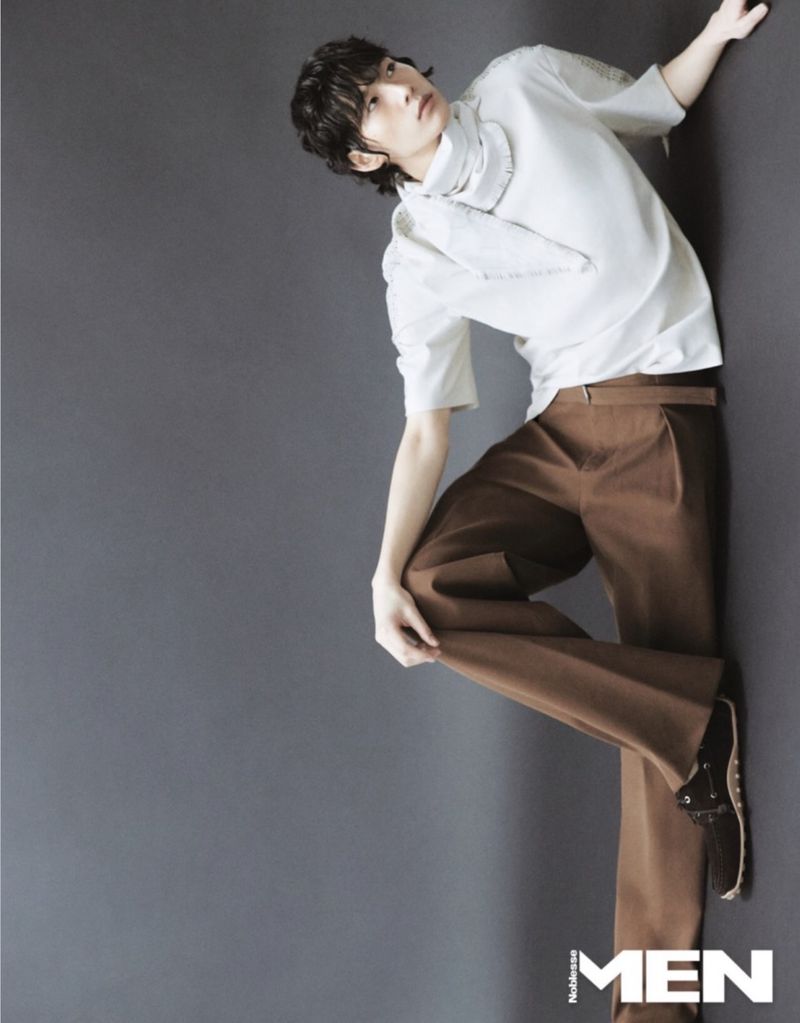 Stylish male fashion model in a white shirt, brown pants, and black shoes posing against a gray backdrop in a studio setting.