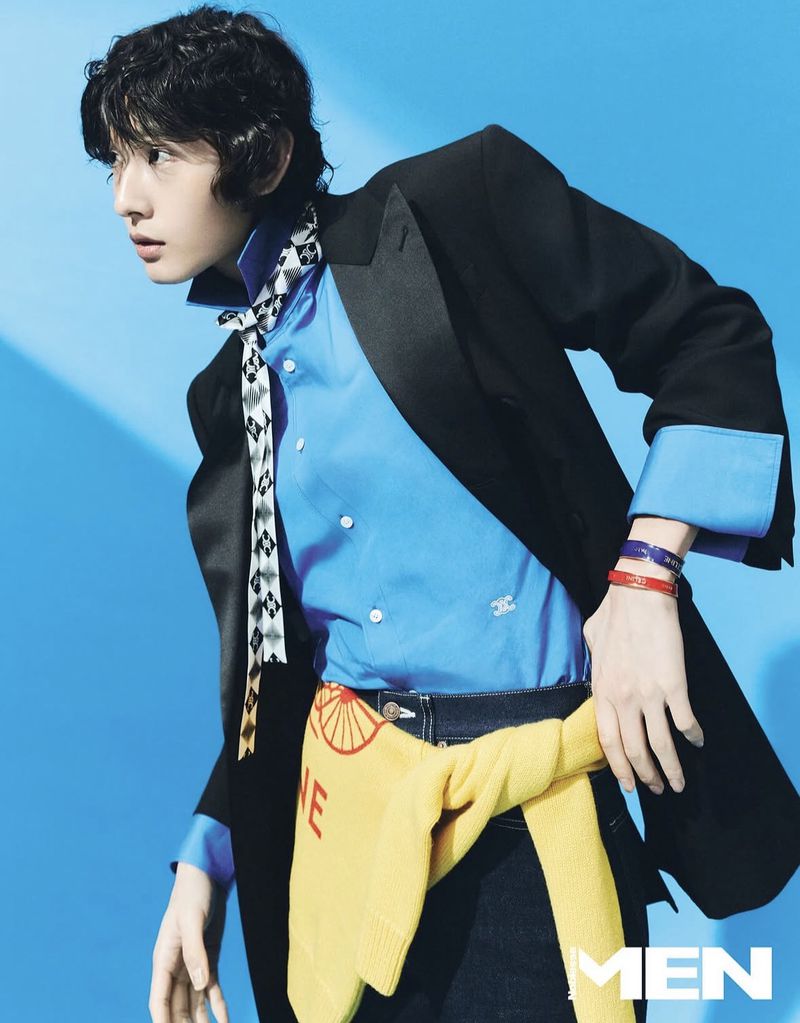 Portrait of a stylish Asian male model in a black blazer, blue shirt, and dark jeans against a vibrant blue backdrop in a studio setting.