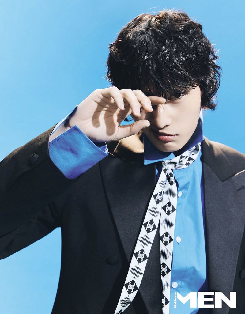 Model with dark curly hair wearing a dark blazer over a blue shirt and patterned tie against a bright blue background