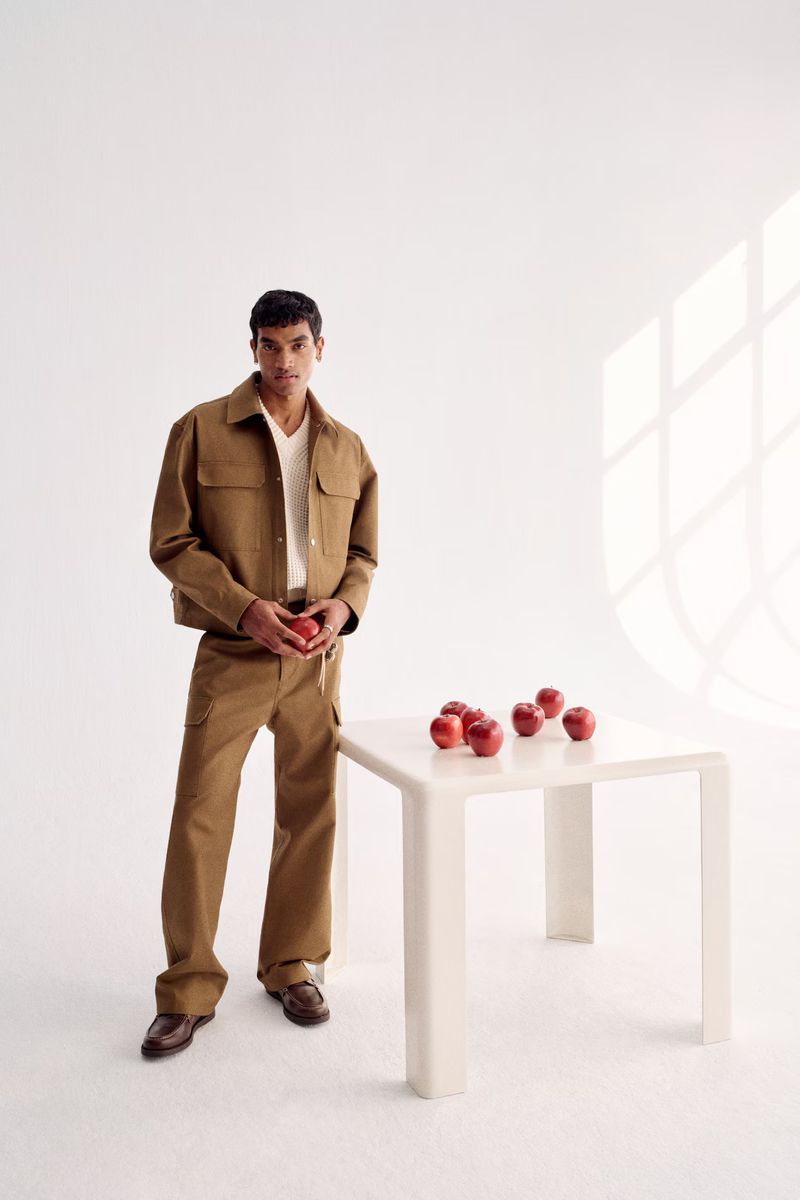 A man in a brown jacket and pants stands indoors with apples on a white table, bathed in natural light creating a minimalist fashion shot.