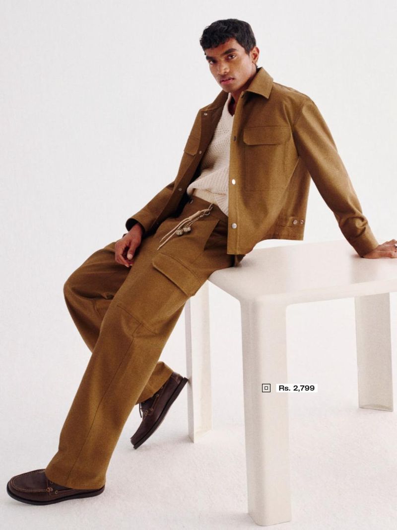 A dark-skinned man in a brown corduroy jacket and trousers poses while sitting on a white table in a brightly lit studio.