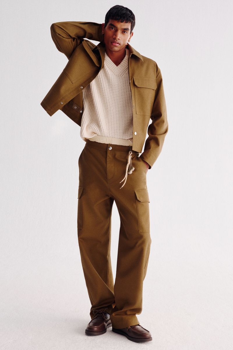 Male model wearing an olive-green jacket and wide-leg cargo pants in a studio shot, posing with hand on his head, fashion editorial.