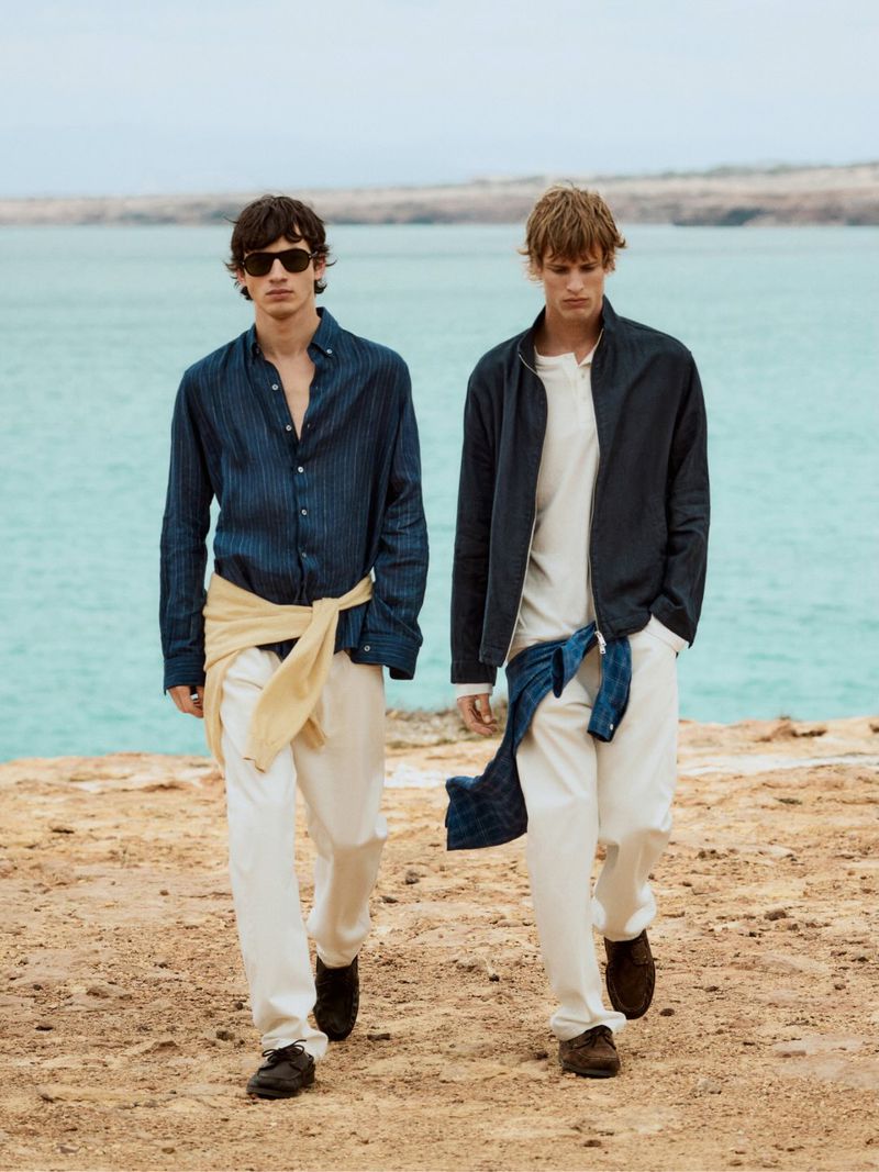 Two adult men walking on a sandy beach, wearing linen pants, button-down shirts and jackets; coastal landscape and ocean background.