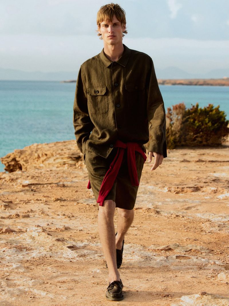 A fair-skinned male model walks on a sandy beach near turquoise water, wearing a dark linen shirt, shorts, and moccasins, with a red sash.