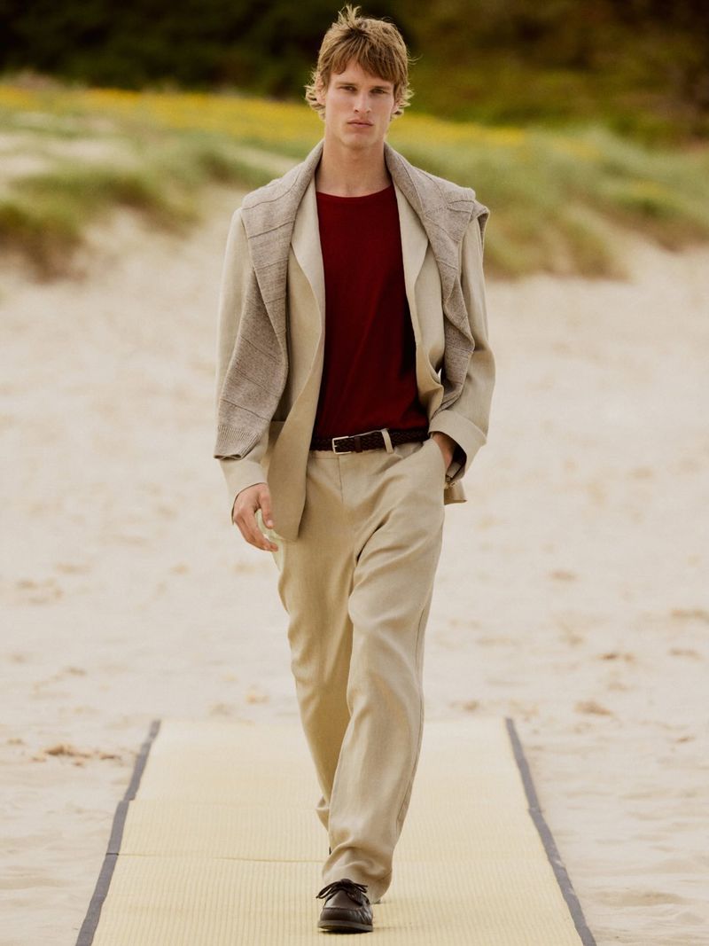 Man in a tan suit walks along a sandy beach on a woven mat. He is wearing a red shirt and has a sweater draped over his shoulders.