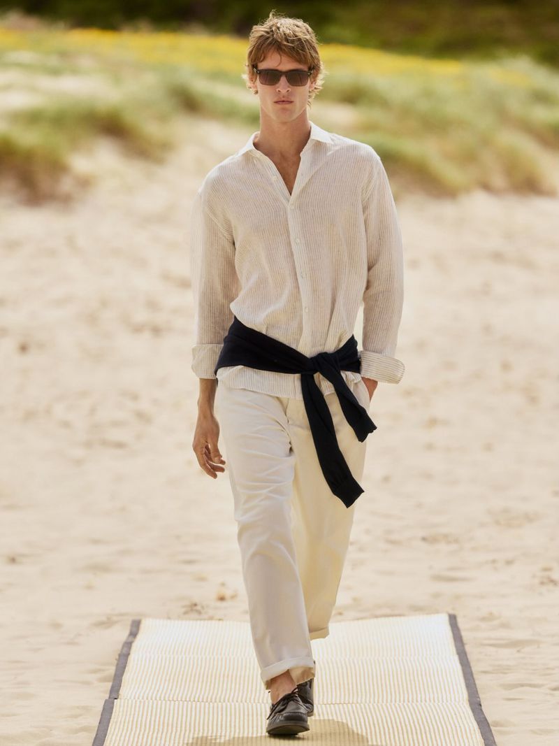 Man in white linen shirt and pants walks on a beach runway with sunglasses. Beach vegetation in background. Fashion and lifestyle shoot.