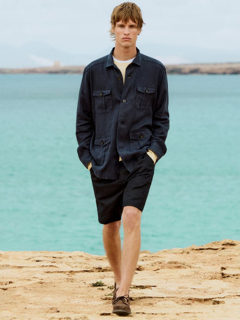 Man in a blue jacket and black shorts walks on a rocky surface near the sea. He wears brown shoes; the sky is overcast.