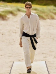 Male model wearing a white linen shirt, trousers, and a dark sweater tied around his waist walking on a sandy beach path