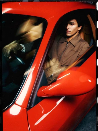 A brunette man is driving a red sportscar, looking out the window with a focused expression in an indoor environment at night.