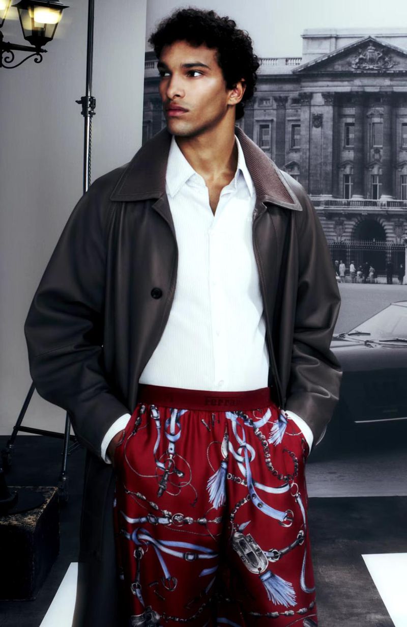 Male fashion model poses in a grey coat and patterned pants in a studio with a city background, showcasing modern elegance and style.