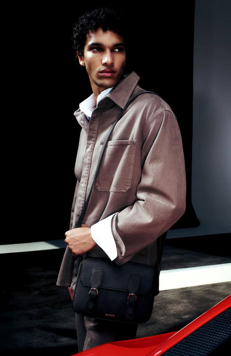 Fashion shot of a young adult male wearing a tan shirt jacket with a black messenger bag against a dark background.