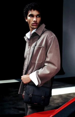 Fashion shot of a young adult male wearing a tan shirt jacket with a black messenger bag against a dark background.