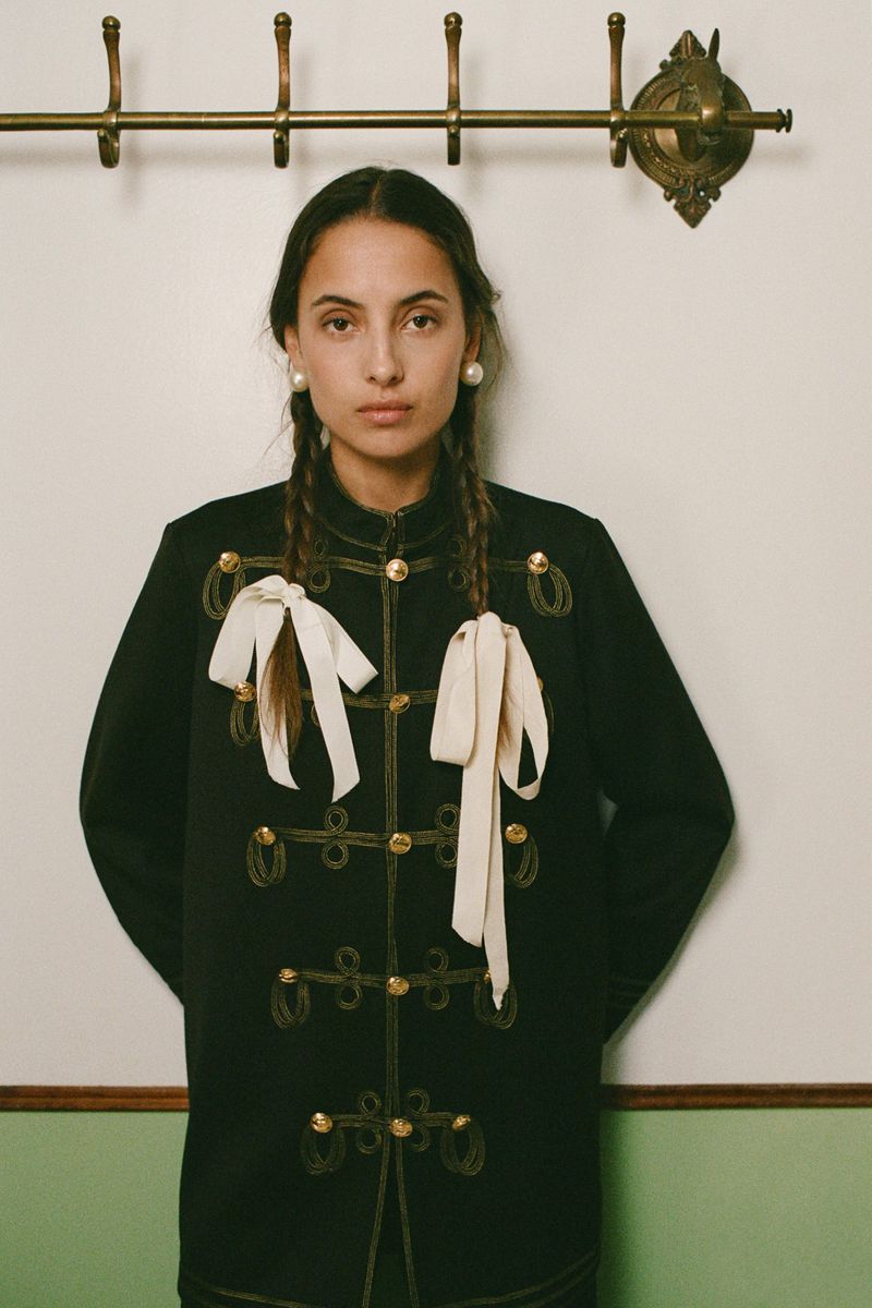 Woman in black jacket with white ribbons and braids stands against a wall with a brass coat rack, portrait shot, studio lighting.