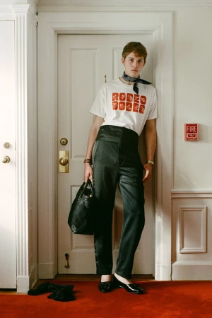 Young adult man poses in a hallway wearing a patterned white t-shirt, black trousers, and black flats holding a black handbag.