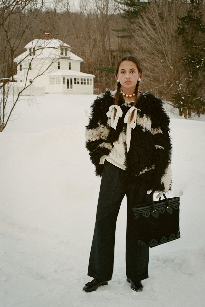 A brunette woman stands in a snowy landscape wearing a fur coat, holding a black bag, with a white house and bare trees behind her.