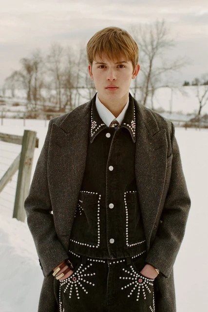 Young man in embellished Western wear and grey coat stands in a snowy rural landscape, facing camera, portraying rugged individualism.