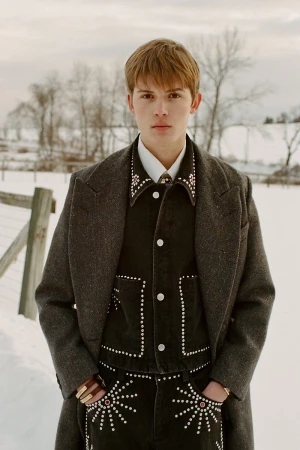 Young man in embellished Western wear and grey coat stands in a snowy rural landscape, facing camera, portraying rugged individualism.