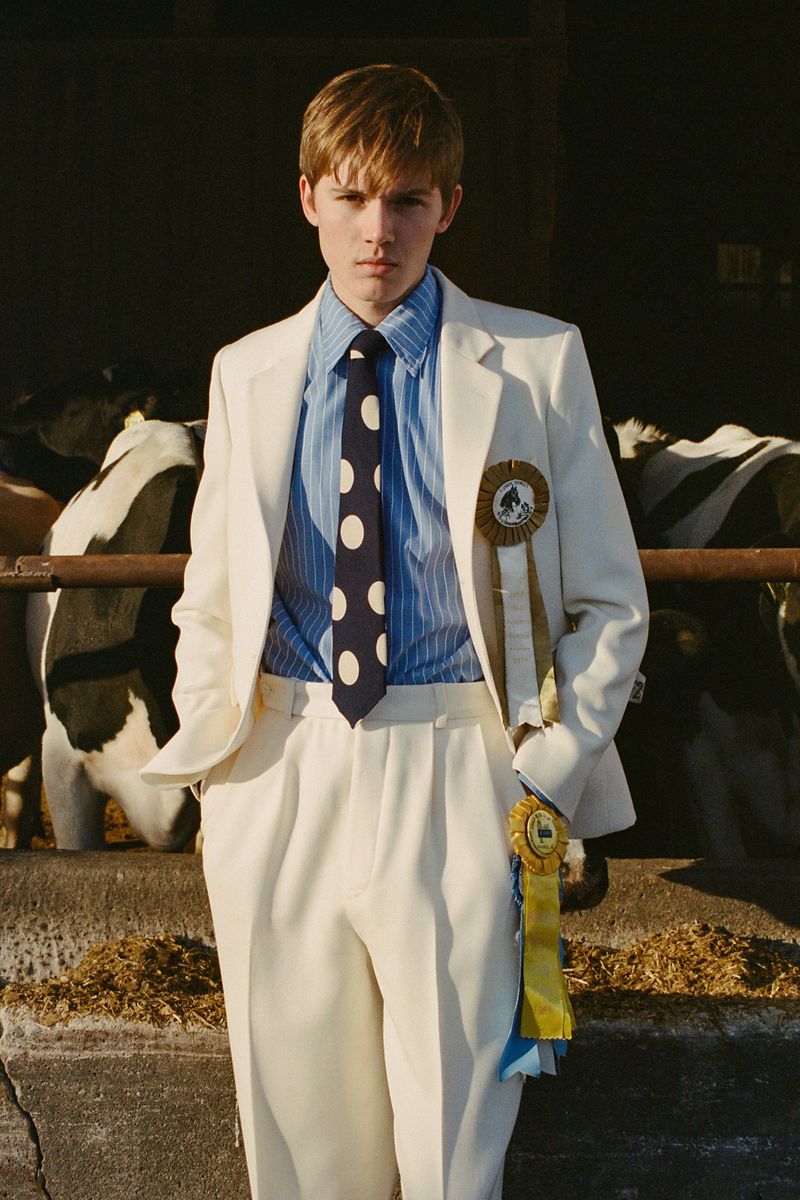 Young man in a white suit and polka-dot tie stands in a barn near cows, wearing an award ribbon, captured in medium shot.